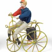 Nr. 104 Radfahrer, männliche Puppe mit Porzellankopf und anmodellierter Kappe auf einem dreirädrigen Fahrrad, H. 25,5 cm Rufpreis € 1.500