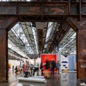 Art Düsseldorf 2023: Besucher erkunden eine Kunstmesse in einer Industriehalle mit rostigen Stahlträgern, modernen Kunstwerken und lichtdurchfluteter Architektur. (c) Foto Sebastian.Drüen