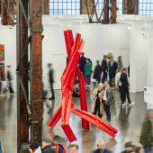 Art Düsseldorf 2023: Besucher flanieren durch die Messe mit moderner Kunst, darunter eine auffällige rote Skulptur vor rostigen Stahlträgern in einer Industriehalle. (c) Foto Sebastian.Drüen