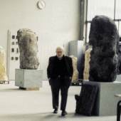 Hans Josephsohn stands in his workshop among monumental sculptures with raw, textured surfaces. The setting reflects his engagement with form and presence.